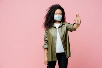 Horizontal shot of Afro American woman keeps palm towards camera, makes stop gesture, tries to prevent coronavirus or Covid-19, wears protective sterile mask, says no to world pandemic disease