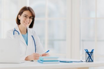 Medical specialist wears white gown with stethoscope, works in hospital, makes notes, poses at own cabinet, sits at table with laptop computer in office smiles happily. Health care and medical concept