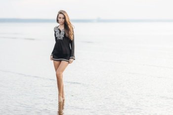 Horizontal shot of pretty female in black blouse, demonstrates her perfect body and slender legs, stands against ocean background, looks with confident expression at camera, poses for magazine