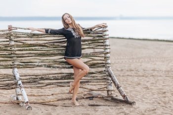 Adorable slim young female model stands on beach near wooden hence, demonstrates slender legs, recreats alone at seaside, feels relaxation and freedom. Summer holidays and relaxation concept
