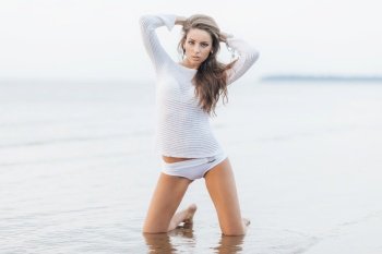 Portrait of attractive female with make up, dressed in white clothes, stands on knees at sandy beach near ocean view, has confident expression, feeles relaxed and free. Woman on coastline alone