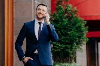 Optimistic male owner of buisness company dressed in elegant suit, has telephone conversation, consults with operator, looks into distance, has happy look, stands outside. People and communication