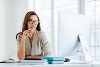 Contemplative female entrepreneur keeps pen in mouth, focused in monitor of computer, thinks on development of new strategy, wears elegant clothes and spectacles, poses against office interior
