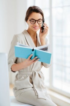 Vertical shot of female administrative manager involved in working process, holds notepad with written notes, discusses something via cellphone, wears spectacles and elegant costume, smiles gently