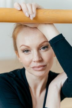 Close up shot of restful woman with ginger hair, has make up, looks at camera, has serious expression, holds hand on handrail, busy doing gymnastic exercises, has natural beauty. Ballet concept