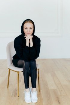 Vertical shot of good looking woman with make up, keeps hands under chin, wears hoody, leggings and white sportshoes, has slender legs, poses on chair in empty room, ready for sport training
