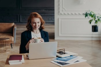 Glad red haired woman has distance job, communicates in video chat with colleagues, discusses ideas for project, enjoys online conferene uses modern laptop and accessories satisfied with friendly talk