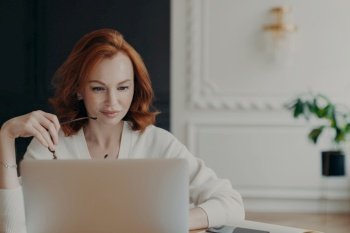 Photo of serious redhead woman journalist concentrated at laptop computer, searches information for article on internet website, holds spectacles, wears white jumper, checks email, works remotely
