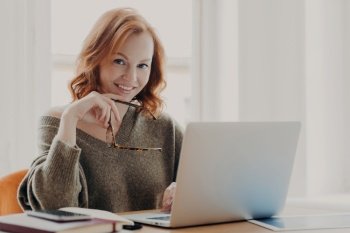 Confident joyful redhead woman works on modern laptop, poses in coworking space, wears sweater, works remotely with application on device, smiles pleasantly, holds spectacles, enjoys distance job