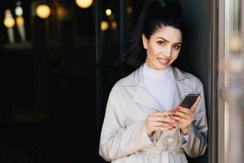 Portrait of adorable bruntte woman with dark eyes and eyebrows, pure healthy skin dressed in white clothes holding mobile phone in hands demonstrating her perfect manicure looking into camera