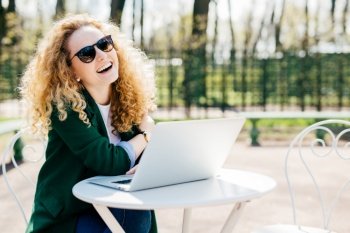 Happy pretty blonde woman with curly hair wearing sunglasses and green jacket having rest in park sitting at white desk using her laptop communicating with friends online. People, emotions concept
