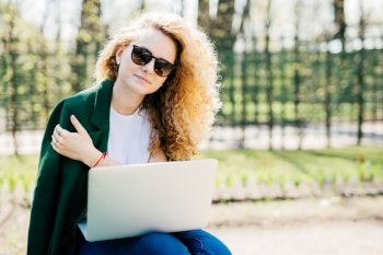 Portrait of cute young woman with curly blonde hair wearing sunglasses, white T-shirt and green jacket holding laptop on knees working at her future project isolated over green nature background