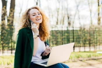 Portrait of beautiful girl having fluffy blonde hair holding smartphone in one hand and sunglasses in other having telephone conversation being excited to speak with her best friend using laptop