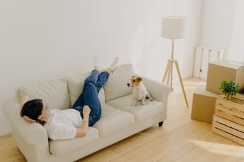 Relocation and mortgage concept. Relaxed brunette woman has rest on comfortable sofa, wears t shirt, jeans and white socks, plays with dog, moves in new apartment, carton boxes with stuff near