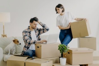 Unhappy male freelancer in spectacles sits in front of modern laptop, works remotely at home, busy woman stands near with carton box, move in new apartment, unpack belongings, pose in living room