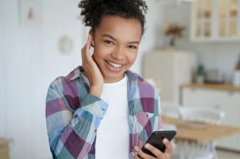 Smiling young mixed race teen girl listens to music in wireless earphone holding smartphone, using mobile apps at home. Happy biracial teen lady enjoy sound, listening to audio book or podcast indoors. Smiling mixed race girl listens to music in earphone holding smartphone, using mobile apps at home