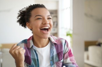 Excited mixed race teen girl makes yes winner hand gesture, celebrates success, personal achievement at home. Overjoyed young lady feeling happy received good news, screaming with raised clenched fist. Excited mixed race teen girl makes yes winner hand gesture, celebrates success, personal achievement