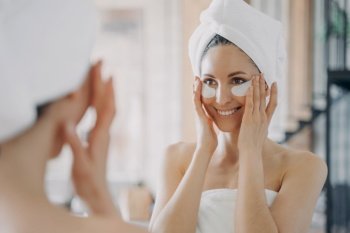 Attractive hispanic woman with eye patches looking at mirror during morning skincare routine. Smiling pretty female wrapped in towel after shower reducing facial wrinkles moisturizing skin under eyes.. Attractive latina woman with under eye patches looking at mirror during daily skincare treatment
