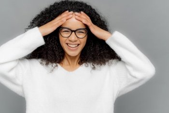 Indoor shot of glad optimistic dark skinned lady touches forehead, has curly Afro hair, giggles positively, watches comedy, wears white sweater, isolated on grey background. Facial expressions