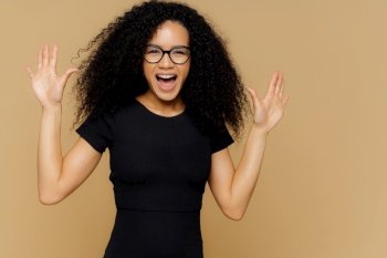 Half length shot of pleased overjoyed woman dances and moves positively, raises palms, exclaims with happiness, wears black t shirt, spectacles, isolated on beige wall. Excited female models moves