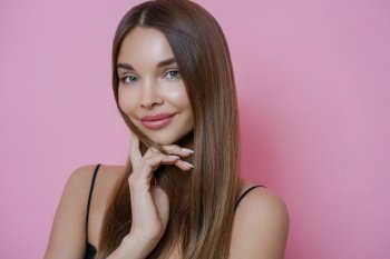 Portrait of attractive woman touches chin gently, has straight hair, stands with naked shoulders, smiles pleasantly, isolated on pink background. Women natural beauty, wellness and elegance concept