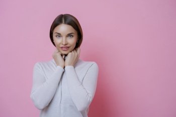 Photo of pleased dark haired woman looks with shy expression at camera, keeps hand under chin, wears white turtleneck, has healthy smooth skin, isolated over pink background, empty space for text