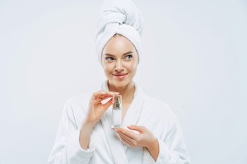 Thoughtful beautiful woman with clean fresh skin, recommends cosmetic product, feels relaxed after taking bath, wears dressing gown, wrapped towel on head. Beauty, wellness, body care concept