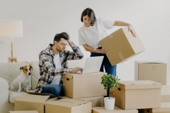Moving concept. Frustrated young man uses modern laptop computer for searching necessary information online, woman replaces big carton boxes with personal belongings. New home and life concept