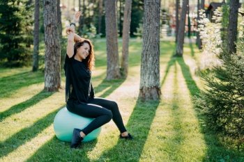 Relaxed brunette woman sits on fitness ball raises arms and does excercises outdoor in forest poses through trees on green grass has athletic body happy expression. Female fitness instructor