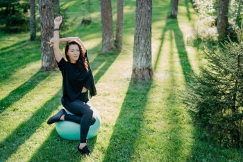Photo of active flexible brunette woman stretches with fitball, does pilates or fitness exercises, poses on green grass during sunny spring day in forest, enjoys gymnastic exercises on fresh air.