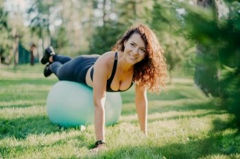 Happy active sportswoman does fitness exercises with gymnastic ball, dressed in active wear, has workout in open air, poses on green grass in forest or park, focused somewhere with pleasant smile
