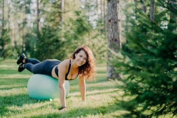 Active lifestyle fitness and body care concept. Fit pleased young brunette woman wears activewear exercises with fitness ball poses in forest on green grass. Sports people. Pilates exercising