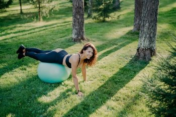 Young pleased sporty woman does fitness exercises with fitness ball, practices yoga in green park or forest, being aerobics instructor, keeps healthy and fit. Physical activity and gymnastics.