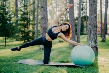 Outdoor shot of active brunette woman in sportswear poses on yoga mat, does stretching exercises with gymnsatic ball, poses in forest or park on green grass. Aerobics, healthy lifestyle concept