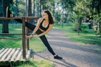 Flexible young curly woman wears black top leggings and sneakers, stretches legs, does sport during summer day in open air, poses against green trees. People, flexibility and exercising concept