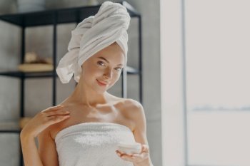 Photo of satisfied young woman has healthy pure skin, applies body cream, uses skin treatment to moisturize, stands wrapped in towel, smiles gently. Personal care, beauty and wellness concept