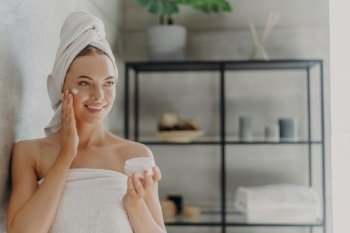 Photo of pretty young European woman applies moisturizing cream on face, has happy expression, wrapped in bath towel, stands against bathroom background indoor. Skin care and hygiene concept
