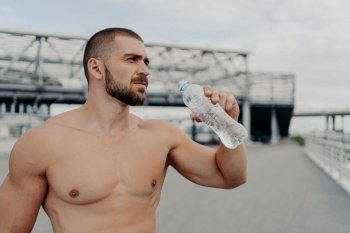 Half length shot of muscular guy drinks fresh water from bottle, takes rest after jogging exercising, has muscular arms, looks into distance thoughtfully, poses outside, cares about water balance