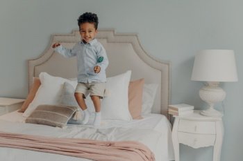 Happy little energetic afro american kid boy jumping alone on his parents bed while holding delicious lollipop, eating sweets, causing mess while no one is home, cute child having fun at home. Energetic afro american kid jumping with lollipop on bed mattress