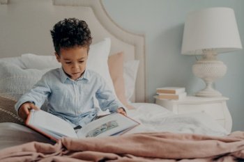 Cute focused small boy in casual wear holds and tries to read childrens book, sitting on bed with nightstand next to with white elegant lamp on it. Kid spending weekend morning leisure at home. Cute afro american small boy trying to read children’s book
