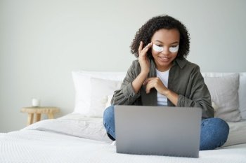 Pretty african american young girl applying under eye patches on face sitting at laptop on bed. Smiling lady with under-eye masks watching series on computer in bedroom. Skincare beauty routine.. African american girl applying under eye patches sitting at laptop on bed. Skincare beauty routine