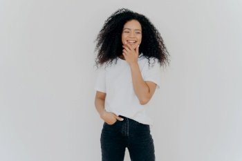Overjoyed mixed race curly woman being in good mood, holds chin and giggles happily, keeps hand in pocket, dressed in casul wear, poses against white studio wall, has fun, hears pleasant news