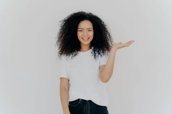 Studio shot of cheerful African American woman grins happily, keeps palm raised over copy space, pretends holding something, wears white t shirt, present good product, recommends nice choice
