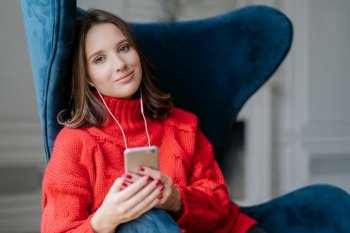 Pretty female teenager enjoys free time and popular music in earphones, holds modern smart phone, dressed in red knitted sweater, sits in comfortable armchair, uses application, checks new updates