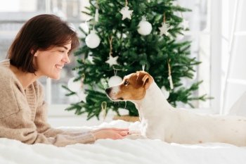 Horizontal shot of beautiful cheerful woman enjoys togetherness with dog, holds its paws, lie on white bed, look each other in eyes, feel love and friendship, decorated Christmas tree in background