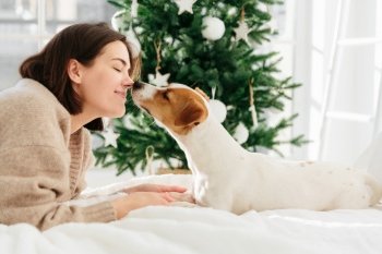Cheerful woman gets kiss from favourite dog, keeps eyes closed from pleasure, lie on bed against Christmas tree at home, have nice relationship. People, New Year, celebration and pets concept