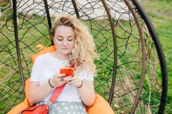 Horizontal shot of pretty young female rests in hanging chair in garden, uses smart phone, being always in touch, checks her email box, dressed casually, has good rest outdoor. Technology concept