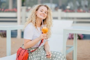 Happy delighted curly young woman dressed in white t shirt and long skirt, holds glass of cocktail, enjoys sunny day and good vacation, has manicure, smiles happily. People and vacation concept