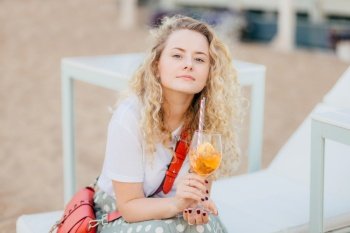 Pleasant looking blonde female has confident expression, looks directly at camera, has pure skin, manicure, holds orange cold cocktail, has pleasant talk with interlocutor, rests outdoor alone