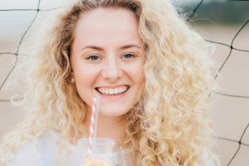 Close up portrait of cheerful young female with curly hair, smiles positively, holds cold drink in front, stands against net background, being in good mood after date with boyfriend. Beauty concept